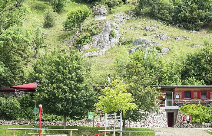 Grüne Hügellandschaft mit Felsen, Bäumen und einem Gebäude im Vordergrund. Spielplatz mit Klettergerüst sichtbar.