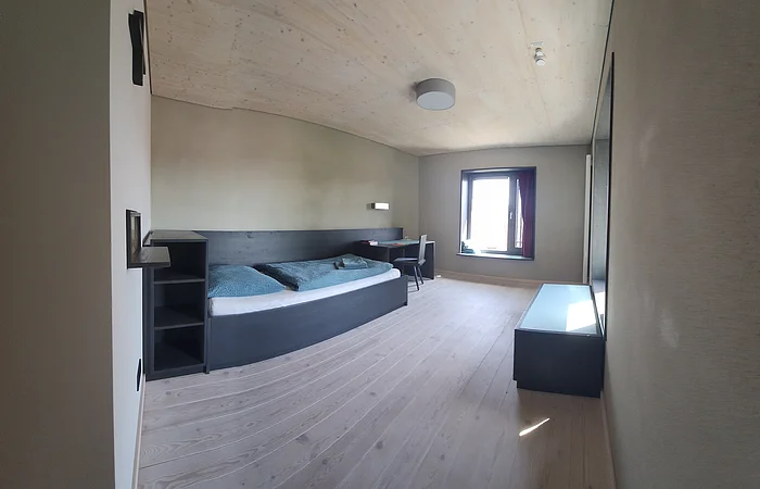 Schlicht eingerichtetes Zimmer mit Einzelbett, Schreibtisch, Stuhl, Fenster und Holzboden.