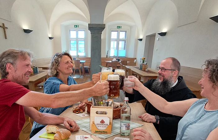 Vier Personen stoßen mit Biergläsern an einem Holztisch in einem hellen Raum mit Kreuz an der Wand an.
