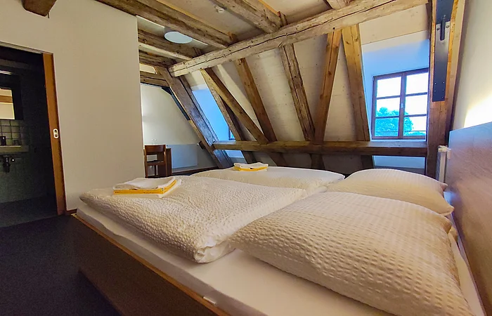 Schlafzimmer mit Dachschräge, Holzbalken, zwei Betten mit weißen Decken und Kissen, Fenster und offenem Badezimmer.