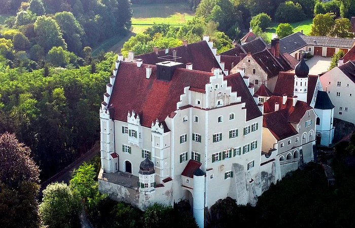 Luftaufnahme eines historischen Schlosses mit roten Dächern, weißen Mauern und umliegendem Wald.
