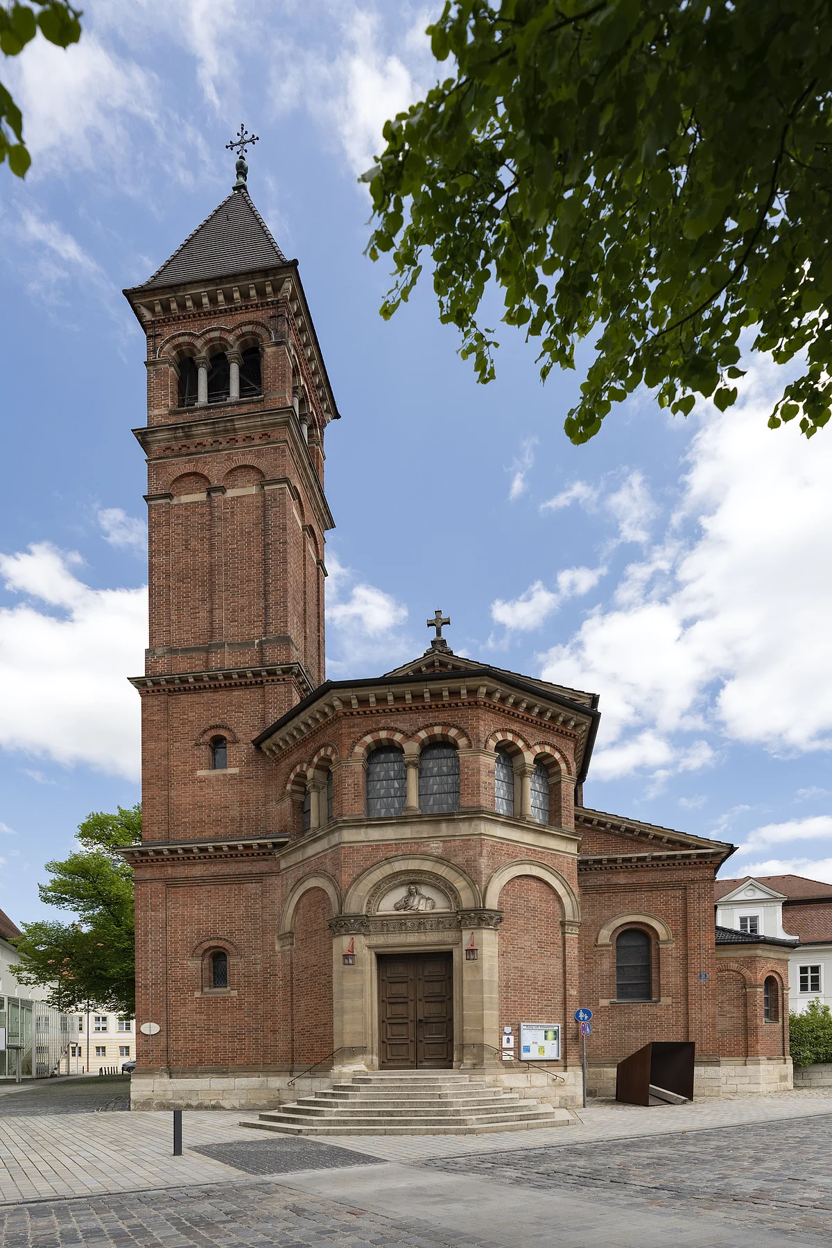 Erlöserkirche (Außenansicht) Das Bild zeigt eine Kirche aus roten Ziegeln. In der Mitte führt eine helle Steintreppe zum Kircheneingang. Über dem Portal befindet sich ein Abbild von Jesus. Auf der linken Seite der Kirche erhebt sich ein Glockenturm mit schwarzem Dach, der fast doppelt so hoch wie die Kirche ist. Darauf steht ein Kreuz.