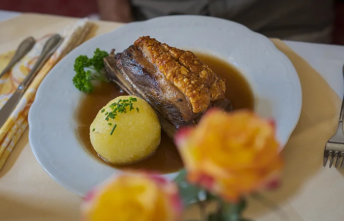 Teller mit Schweinebraten, Knödel und Soße, Besteck und Serviette auf gedecktem Tisch, unscharfe Blumen im Vordergrund.