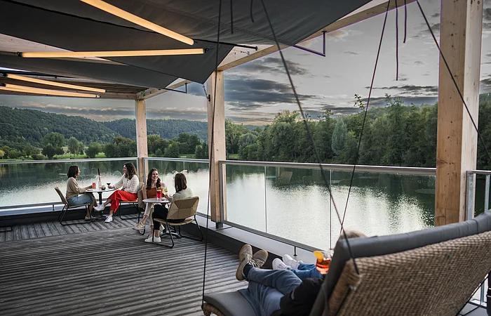 Vier Frauen sitzen an zwei Tischen auf einer Terrasse mit Blick auf einen See und bewaldete Hügel.