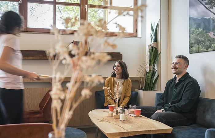 Zwei Personen sitzen an einem Holztisch in einem Café, eine Bedienung bringt Getränke, Pflanzen und Fenster im Hintergrund.