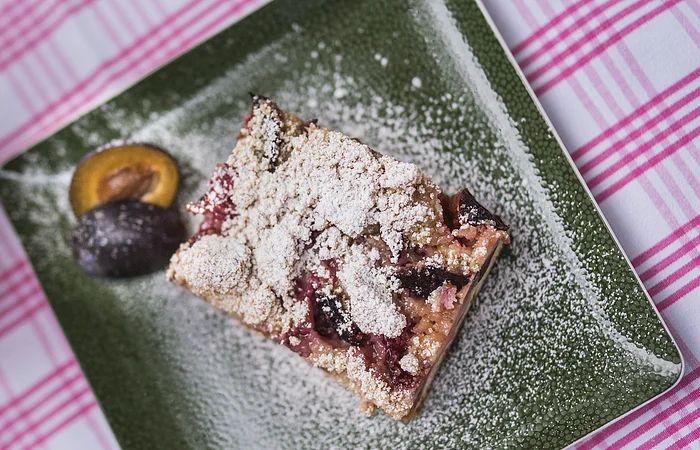 Stück Pflaumenkuchen mit Puderzucker auf grünem Teller, daneben halbierte Pflaumen, auf kariertem Tischtuch.