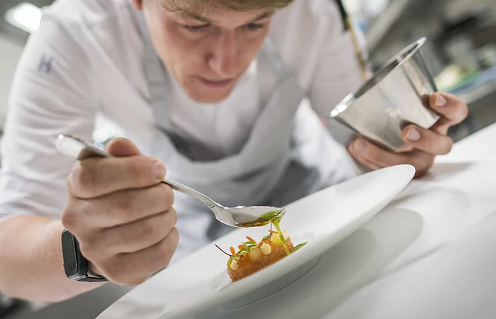 Koch trägt weiße Kleidung und gibt mit einem Löffel grüne Sauce auf ein angerichtetes Gericht auf weißem Teller.