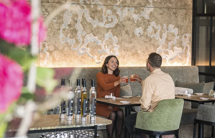 Zwei Personen sitzen in einem Restaurant und stoßen mit Getränken an, im Vordergrund unscharfe Blumen und Flaschen.