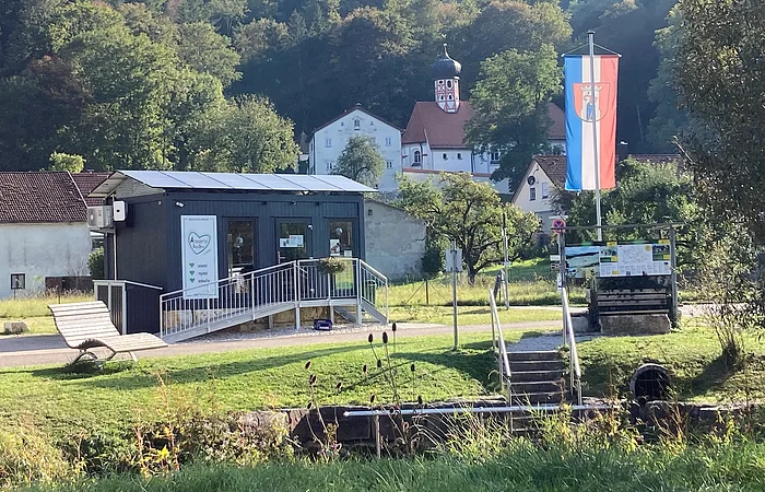 Kleines Gebäude mit Rampe, Bank, Fahne und Kirche im Hintergrund vor bewaldetem Hügel bei Sonnenschein.