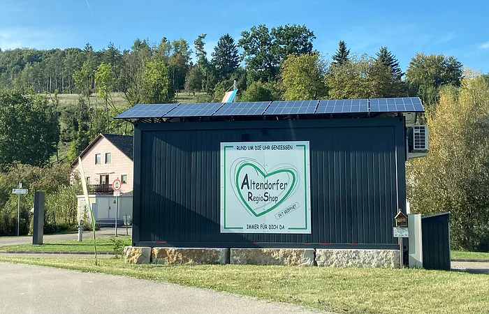 Schwarzer Verkaufscontainer mit Solarpanelen und Schild „Altendorfer RegioShop“ vor Wald und Haus bei blauem Himmel.