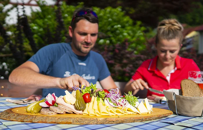 Zwei Gäste genießen üppige Brotzeitplatte im Gasthof "Zum blauen Hecht" in Kipfenberg. Die Platte ist bestückt mit verschiedenen Käse- und Wurstaufschnitten, Radieschen, Tomaten, Essigkuren und roten Zwiebeln. Daneben steht ein Brotkorb.