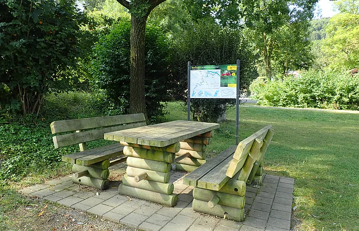 Holztisch mit zwei Bänken auf gepflastertem Platz unter Baum, im Hintergrund Infotafel und Grünfläche.