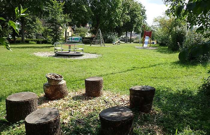 Spielplatz mit Baumstamm-Sitzkreis, Karussell, Klettergerüst und Rutsche auf einer Wiese mit Bäumen im Hintergrund