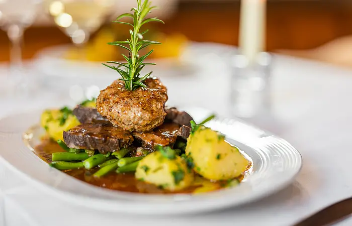 Das Bild zeigt einen Braten, welcher mit viel Hingebung angerichtet wurde. Drappiert ist der Braten auf einem Bett aus grünen Bohnen mit frischen Petersilien-Kartoffeln. Oben auf ein Semmelknödel garniert mit einem Rosmarin-Zweig und schöner Bratensoße