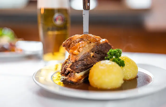 Traditionelles Schweineschäuferl, angerichtet auf einem weißen Teller mit viel Soße, zwei Kartoffelknödel und einem scharfen Messer, welches traditionell senkrecht in das Schäuferl gesteckt wird