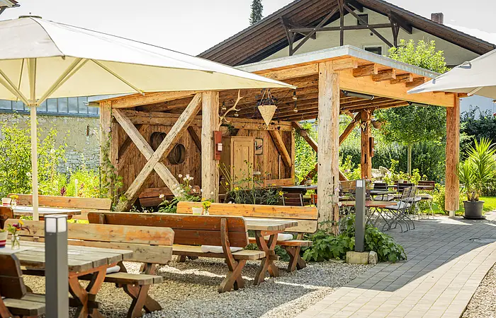 Ein liebevoll und grün gestalteter Biergarten mit Holz-Biertischen, großen weißen Sonnenschirmen sowie einem massiven Holzunterstand, der mit alten Küchenutensilien und Lichterketten dekoriert ist