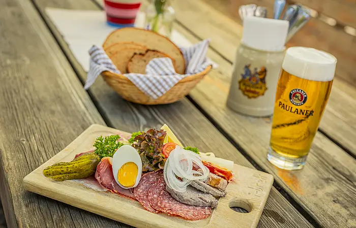 Auf einem Holz-Biertisch steht ein frisch gezapftes Glas helles Bier, ein Brotkorb mit bayerischer Serviette und frischem Brot drin sowie eine liebevoll und typisch bayerische Brotzeitplatte mit frischer Wurst, garniert mit Zwiebeln, Essiggurken und Ei