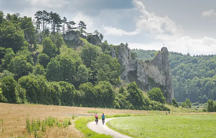 Zwei Radfahrer auf einem Weg vor bewaldeten Felsen mit einem Kreuz auf dem höchsten Felsen.