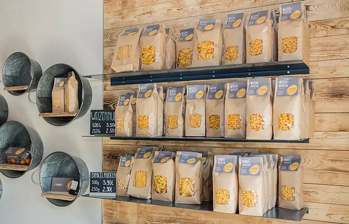Rechts sind drei Regalbretter an einer Holzwand angebracht. Darauf stehen mehrere Nudelpackungen. Daneben an einer weißen Wand sind verschieden große Zinswannen mit Innenbrett als Regale angebracht, in denen sich Regionale Produkte wie Eierlikör, Eier und Nudelpackungen befinden.
