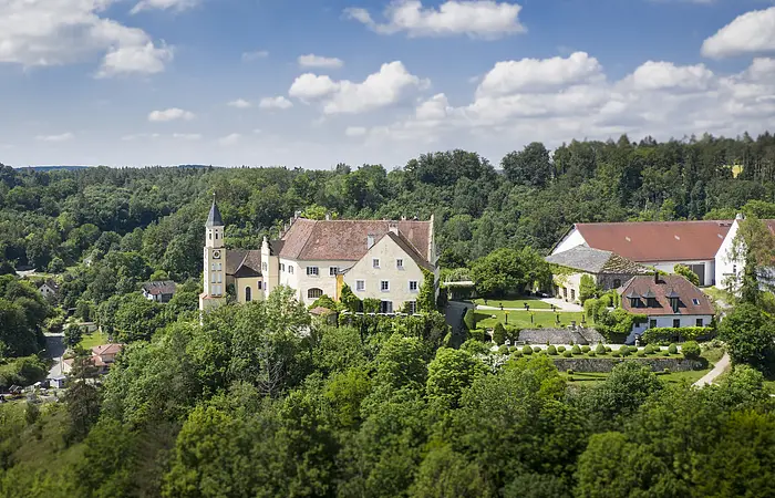 Das Schloss Hexenagger von oben zu sehen. Eingebettet in grünem Blätterkleid. Der Himmel ist hellblau und weiße Wolken sind zu sehen.