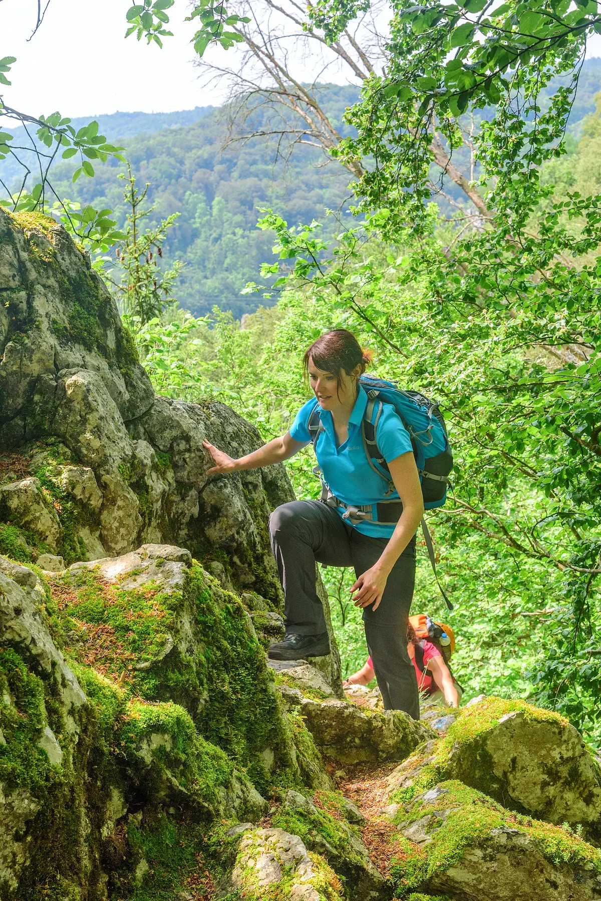 Kletterer am Oberlandsteig Eine Wanderin mit blauem Shirt und Rucksack wandert entlang Felsen, die mit Moos bewachsen hinauf. Weiter unten ist eine zweite Frau in rosa zu sehen. Hinter ihnen ein schöner Blick ins Tal umgeben von grünem Blätterwerk.