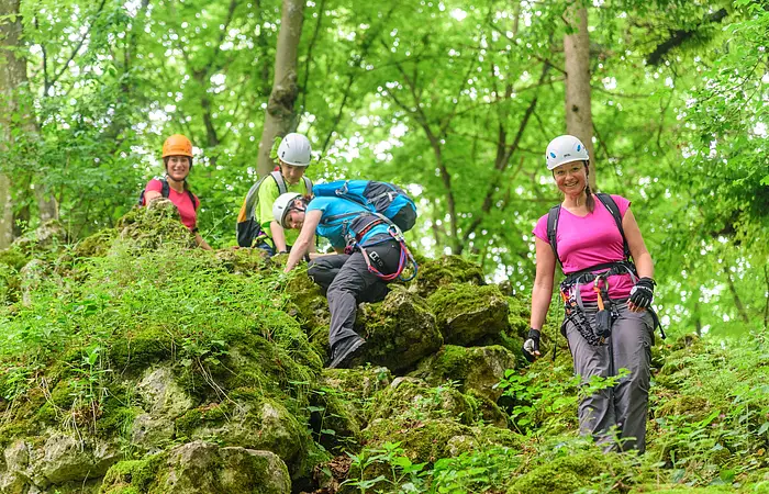 Vier Kletterer mit Helm und Ausrüstung für den Klettersteig gehen einen Hang im Grünen hinunter.