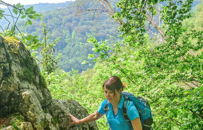 Eine Wanderin mit blauem Shirt und Rucksack wandert entlang Felsen, die mit Moos bewachsen hinauf. Weiter unten ist eine zweite Frau in rosa zu sehen. Hinter ihnen ein schöner Blick ins Tal umgeben von grünem Blätterwerk.