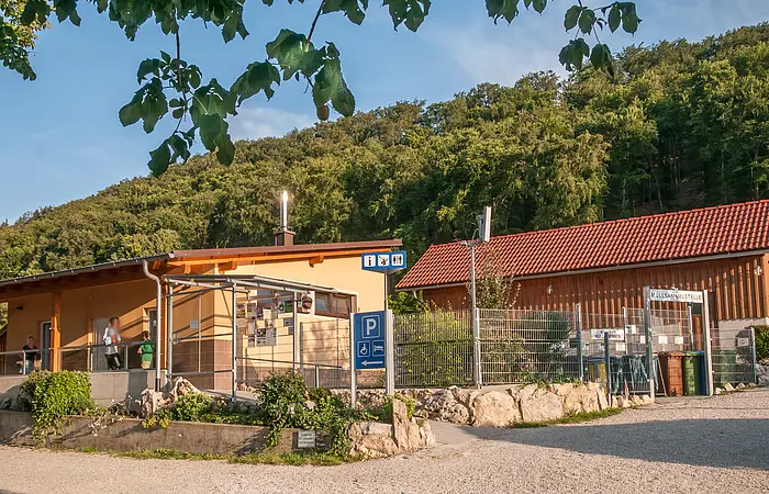 Das Gebäude aus Holz ist l-förmig gebaut und schön mit Pflanzen und Steinen angepflanzt. Hinter dem Campingplatz sieht man den Wald. Der Himmel strahlt himmelblau.