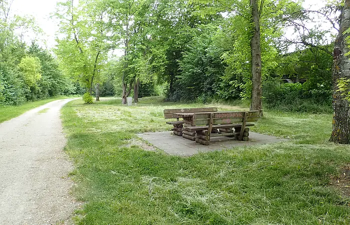 Unter dem Bäumen steht eine gemütliche Sitzgelegenheit aus Holz. Links führt ein geschotterter Weg entlang.