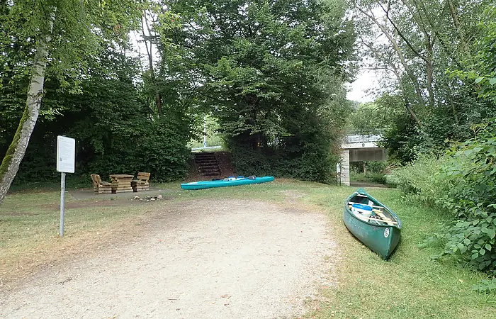 Grüner Kanu und Kajak auf Grasfläche neben Waldweg mit Sitzgruppe und Brücke im Hintergrund.