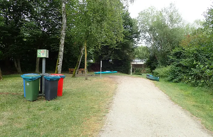 Waldweg mit Mülltonnen, Bänken, Kanus und Brücke im Hintergrund, umgeben von Bäumen und Grasfläche.