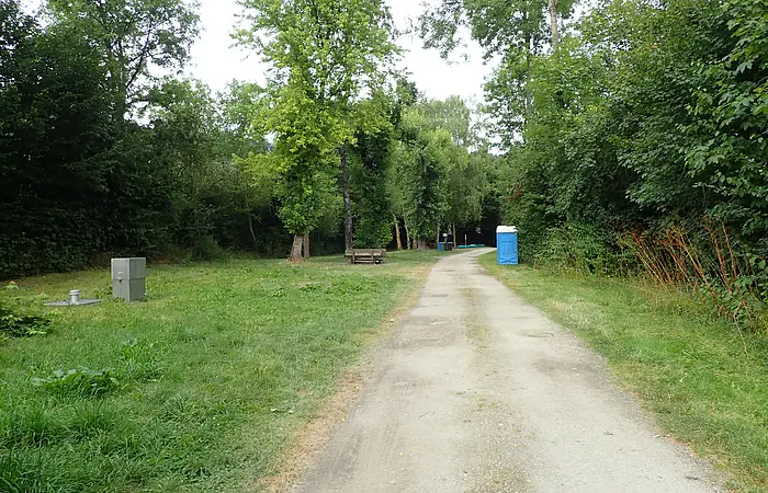 Waldweg mit Grasflächen, Bäumen, einer Bank und einem blauen mobilen Toilettenhäuschen am rechten Wegesrand.