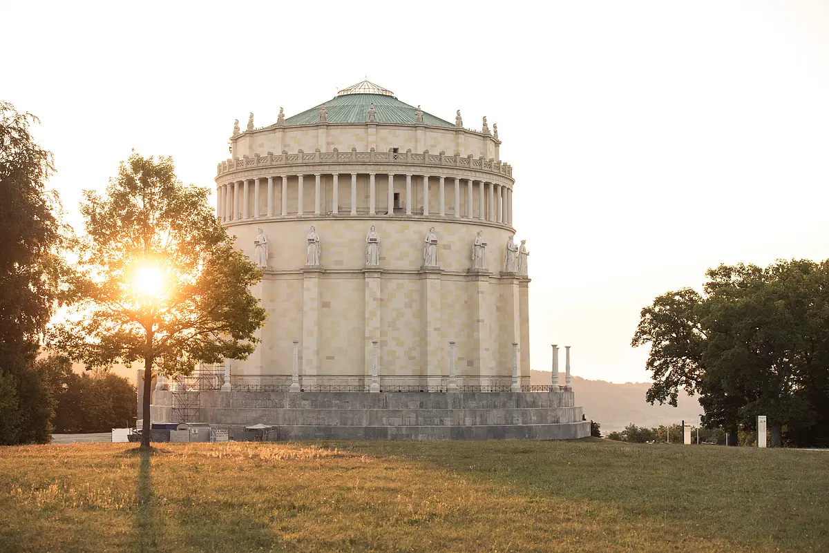 Befreiungshalle in Kelheim von Fotograf Anton Mirwald Rundes historisches Gebäude mit Statuen auf dem Sims, Sonne hinter Baum im Park bei Abendlicht