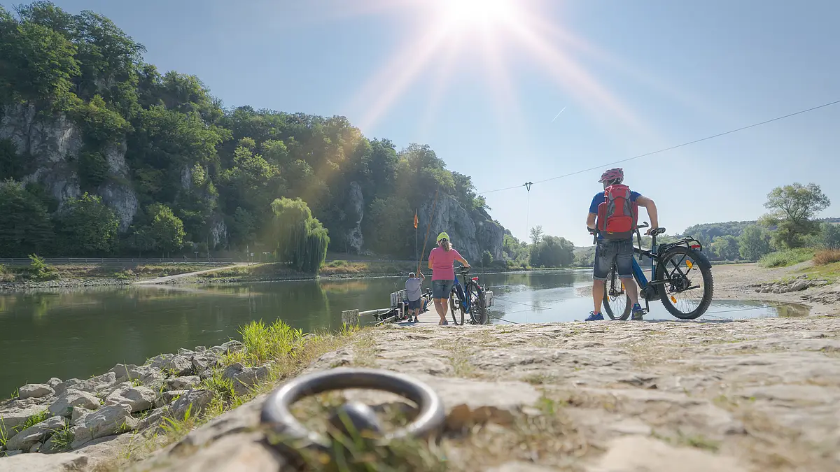 Personen mit Fahrrädern stehen an einem Flussufer mit bewaldeten Felsen im Hintergrund bei Sonnenschein.