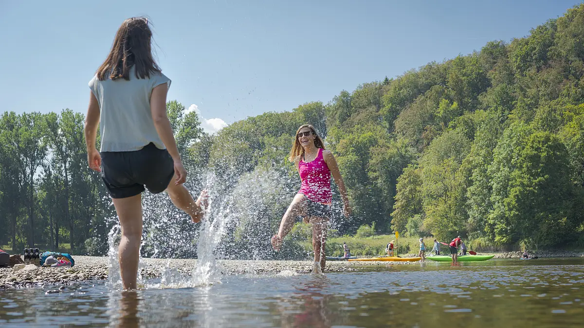 Weltenburger Enge Am Kiesstrand des Klosters Weltenburg stehen zwei Frau und ein paar Meter entfernt ihre Tochter mit den Füßen in der Donau. Beide spritzen sich mit den Füßen nass. Im Hintergrund sind weitere Menschen zu sehen und der grüne Wald der sich am anderen Flußufer erstreckt.
