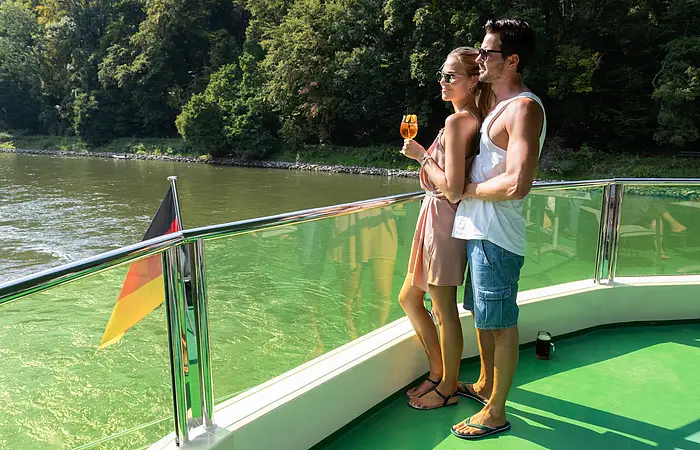 Eine Frau in sommerlicher Kleidung mit Eis in der Hand steht an der Reling. Sie ist an einen Mann hinter hier gelehnt. Beide genießen den Blick aufs Wasser. Eine Deutschlandfahne ist am Heck befestigt.