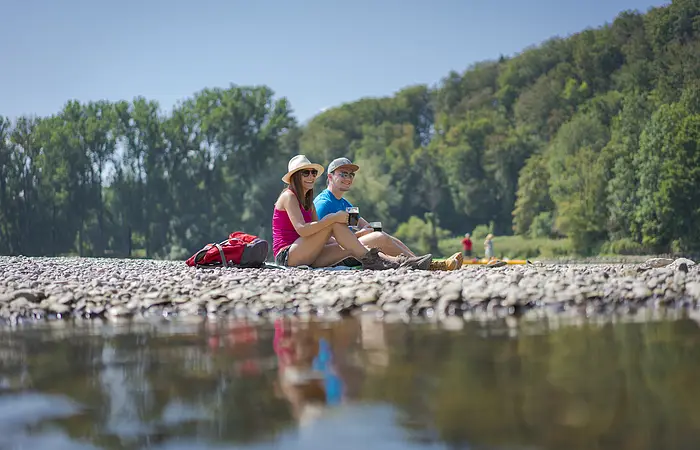 Ein Pärchen mit Hüten sitzt am Kiesstrand des Kloster Weltenburg. Vor ihnen fließt die Donau vorbei und im Hintergrund ist der Wald gegenüber zu sehen.