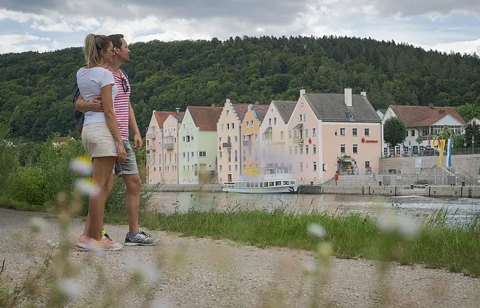 Ein Pärchen in sommerlicher Kleidung steht am Main-Donau-Kanal und halten sich im Arm. Sie schauen aufs Flussufer gegenüber, dort ist die Riedenburger Promenade zu sehen. Dahinter ragen die Hänge mit ihrem dunkelgrünen Kleid auf.