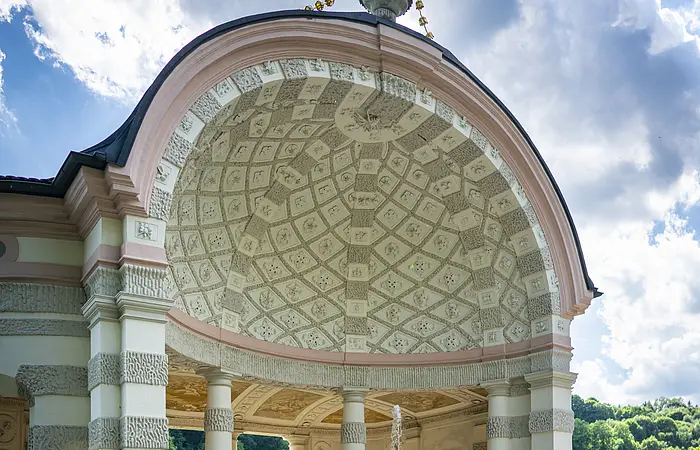 Auf dem Bild ist der mittlere und größte Pavillon des Hofgartens. Der Pavillon ist rund und nach vorne hin geöffnet. Ein Brunnen befindet sich im Zentrum. Dahinter stehen mehrere weiß, grau gemusterte Säulen. Die Kuppel ist innen mit einem mosaikartigen Muster aus grauen und weißen Elementen geschmückt. Das Dach wird von einer vasenähnlichen Figur geziert.
Bildbeschreibung ehemalige Kapuzinerkirche (innen)