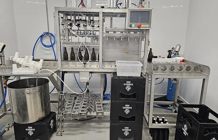 Abfüllanlage mit braunen Flaschen, Metallgestell, Kunststoffkisten und Schläuchen in einem Raum mit weißen Wänden.