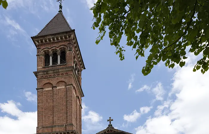 Das Bild zeigt eine Kirche aus roten Ziegeln. In der Mitte führt eine helle Steintreppe zum Kircheneingang. Über dem Portal befindet sich ein Abbild von Jesus. Auf der linken Seite der Kirche erhebt sich ein Glockenturm mit schwarzem Dach, der fast doppelt so hoch wie die Kirche ist. Darauf steht ein Kreuz.