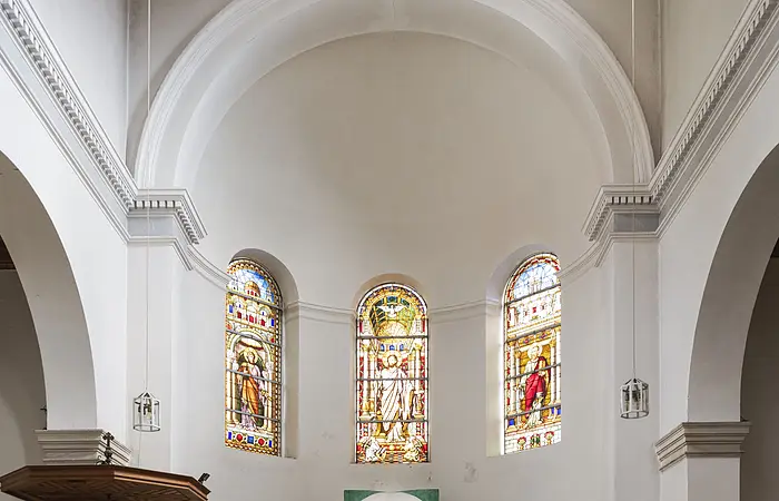 Sie stehen in den Bankreihen der Kirche vor dem Altarraum. Er ist halbrund geformt und die Wände sind weiß. Mittig steht ein großes, modern gestaltetes Kreuz mit Jesusfigur und ein schlichter Altartisch. Darüber sind drei Fenster aus buntem Glas zu sehen, die Petrus, Jesus und Paulus zeigen. Links im Bild befindet sich an einer Säule eine weiße Kanzel mit hölzernem Dach.
Erlöserkirche (Außenansicht)
Das Bild zeigt eine Kirche aus roten Ziegeln. In der Mitte führt eine helle Steintreppe zum Kircheneingang. Über dem Portal befindet sich ein Abbild von Jesus. Auf der linken Seite der Kirche erhebt sich ein Glockenturm mit schwarzem Dach, der fast doppelt so hoch wie die Kirche ist. Darauf steht ein Kreuz.