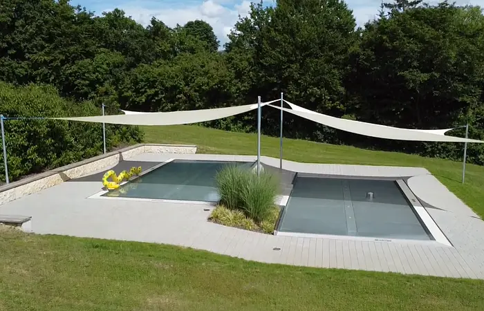 Rechteckiger Pool mit Abdeckung, umgeben von Rasen und Bäumen, mit Sonnensegeln und gelbem Schwimmtier.