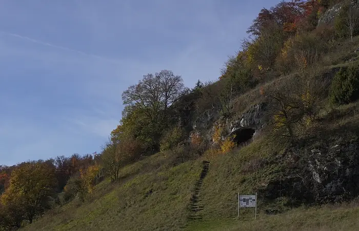 Grasbewachsener Hang mit Treppe, Bäumen und einer Felsöffnung unter blauem Himmel.
