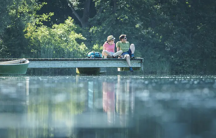 Ein Wanderpärchen sitzt auf dem Steg am Kratzmühlsee und lässt den Blick über den See schweifen. Hinter ihnen die Bäume und links am Steg ist ein Ruderboot befestigt.