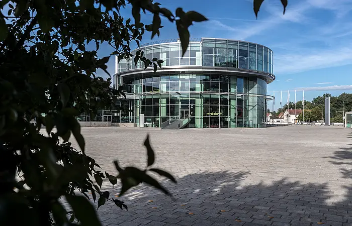 Das Museum mobile mit seiner ungewöhnlichen, runden Architektur. Vor dem Museum erstreckt sich die gepflasterte Piazza und links ragt ein Baum herein.
