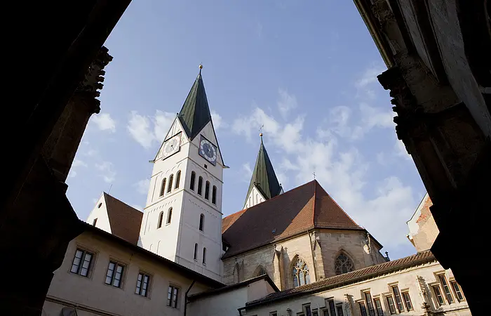 Das Zentrum des Bildes zeigt die Rückseite des Eichstätter Doms. Das Kirchenschiff wird von zwei Kirchtürmen mit jeweils einem dunklen, spitz zulaufenden Dach flankiert. Die Spitzen der Türme bilden jeweils eine goldene Kugel. Das Kirchenschiff ist mit gotischen Fenstern versehen.