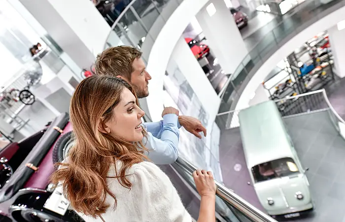 Ein Pärchen entdeckt das Audi Forum. Sie blicken über das Geländer zum unteren Stockwerk wo ausgewählte Fahrzeuge ausgestellt sind.