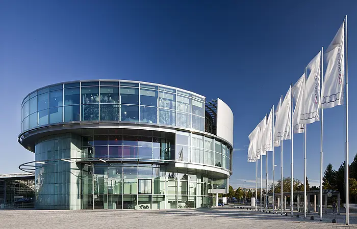 Das Museum mobile mit seiner einzigartigen runden Architektur. Vor dem Museum die Piazza und rechts sind sechs Fahnen gehisst. Der Himmel strahlt wolkenlos.