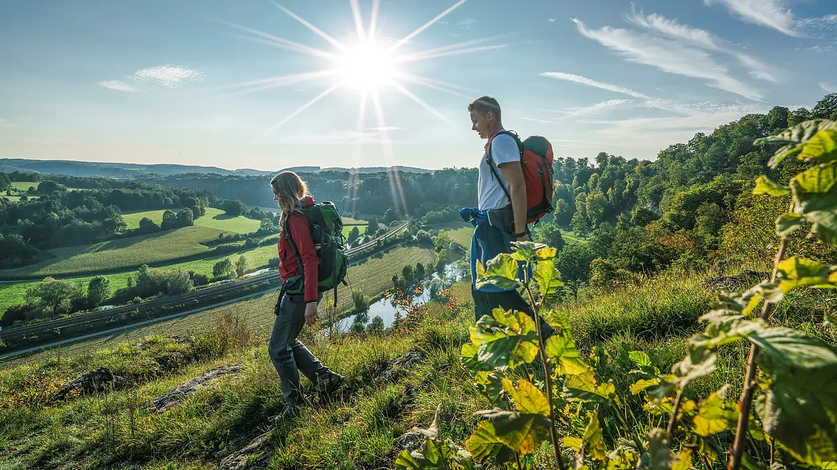 Wanderer oberhalb Hagenacker am Altmühltal-Panoramaweg Eine Frau mit roter Jacke und grünem Rucksack geht vor einem Mann mit rotem Rucksack und weißen Shirt den Altmühltal-Panoramaweg nach Hagenacker hinunter. Die Wiesen sind grün und man hat eine gute Aussicht auf Hackenacker und das Tal.